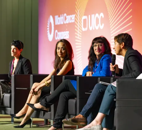 Panel discussion at a conference, with five speakers seated on stage in armchairs, one holding a microphone while speaking. A large screen behind them displays the World Cancer Congress and UICC branding, creating a professional event setting.