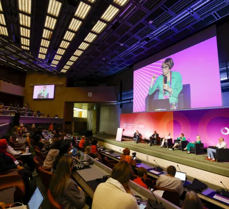 Wide view of a plenary session at the 2024 World Cancer Congress in Geneva, Switzerland, showing a large auditorium filled with attendees seated in tiered rows. On stage, a panel of speakers sit in armchairs against a brightly lit magenta and orange backdrop displaying “World Cancer Congress” and “UICC.” A large screen above the stage shows a close-up of one speaker holding a microphone, while a smaller screen mirrors the same image. The hall features a modern ceiling with grid lighting, and participants in