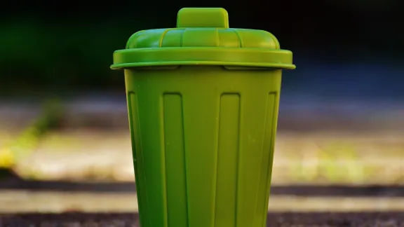 A small green trash can with a lid, featuring vertical ridges, is positioned on a textured surface. The background is blurred, emphasizing the trash can in the foreground.
