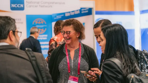 Several World Cancer Congress 2024 participants wearing lanyards stand in conversation near an exhibition booth, with sponsor signage visible in the background.