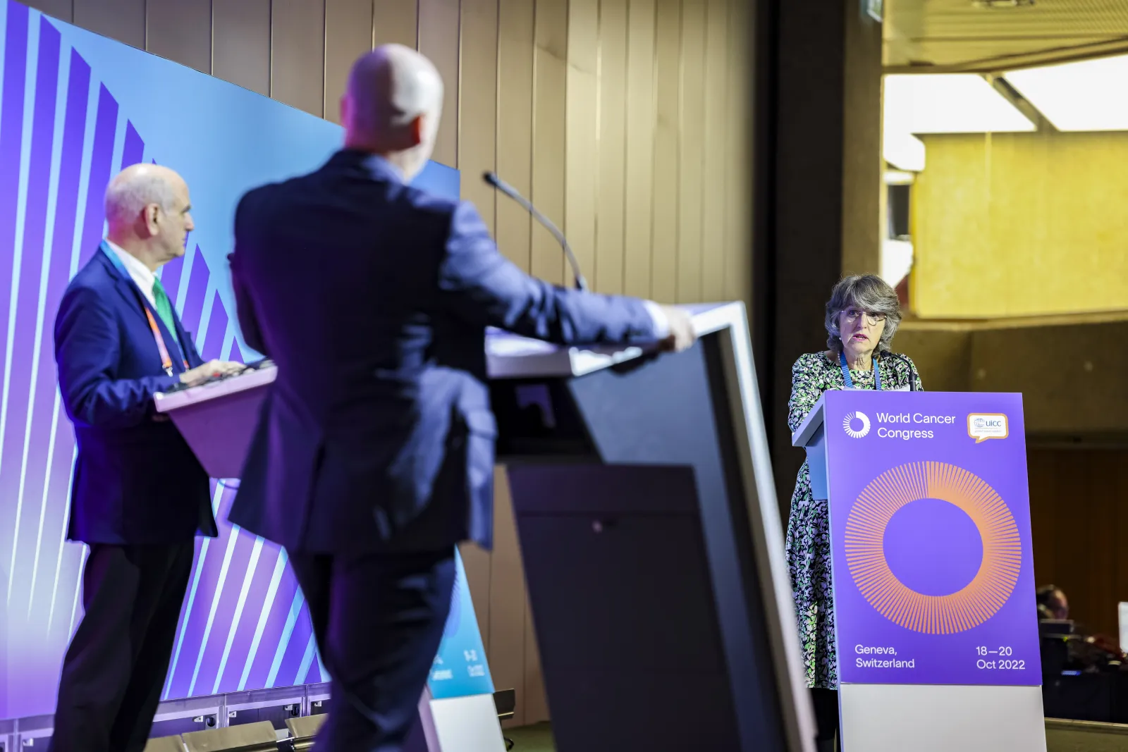 Speaker at a podium during a Big Debate session at the World Cancer Congress 2022 in Geneva, Switzerland, with two additional speakers visible on stage and a branded backdrop displaying the World Cancer Congress logo and event dates.