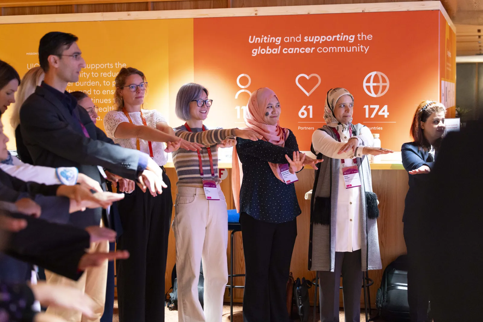 Group activity at the World Cancer Congress 2024 in Geneva, Switzerland, showing participants standing side by side with outstretched arms during an interactive moment in front of a brightly branded UICC backdrop highlighting the global cancer community.