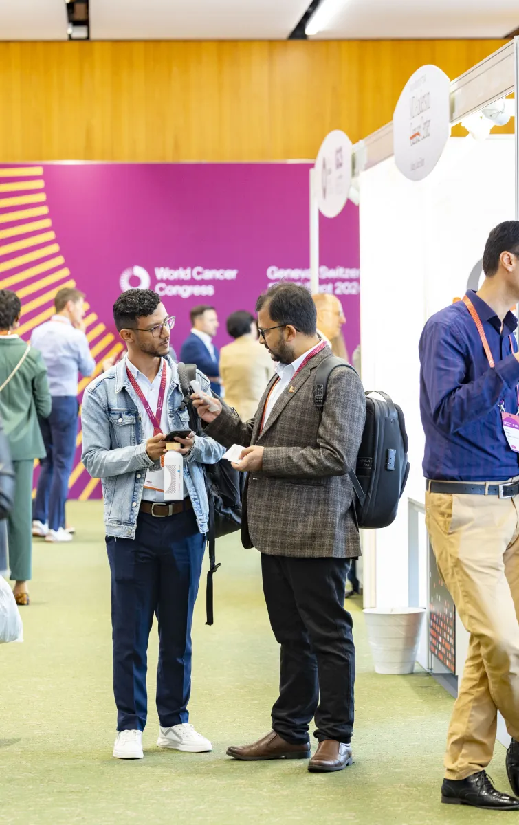 World Cancer Congress 2024 participants wearing lanyards speak together in an exhibition area, with organisational booths and signage visible.