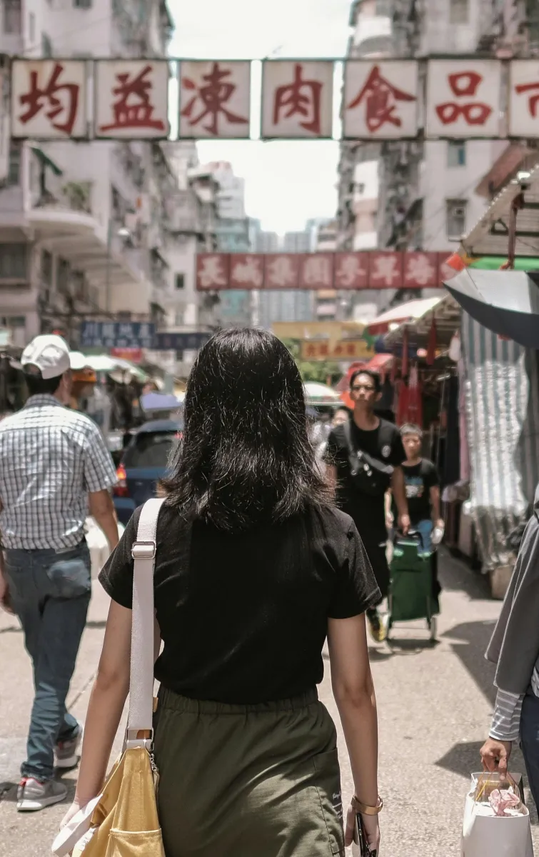 A busy street market in Hong Kong with pedestrians walking between stalls and shops, lined with hanging signage and market umbrellas.