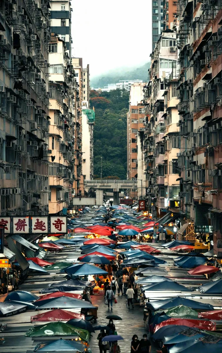 A dense outdoor street market in Hong Kong, with rows of covered stalls and pedestrians between tall residential buildings.